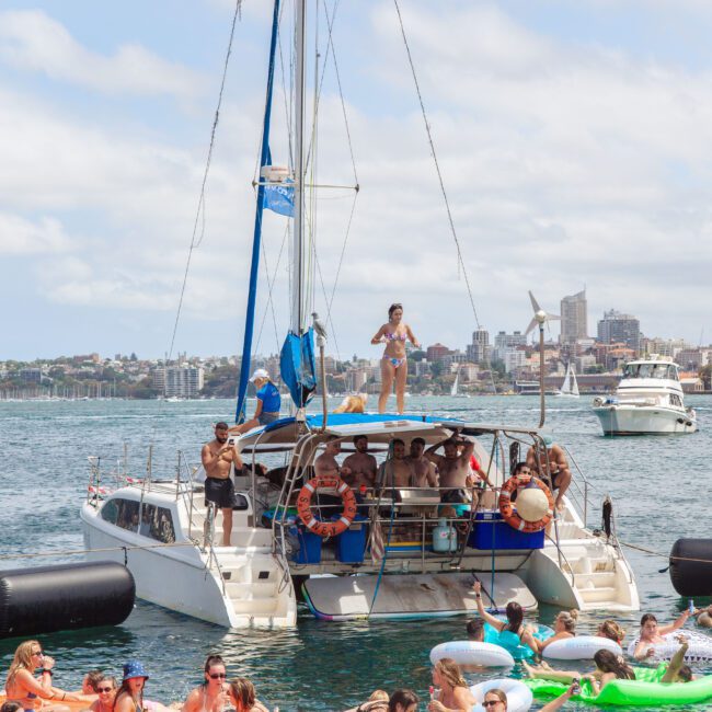 A group of people are gathered on a catamaran and floating nearby in the water, enjoying a sunny day. Some are relaxing on pool floats, others are dancing or standing on the boat, with a city skyline visible in the background.