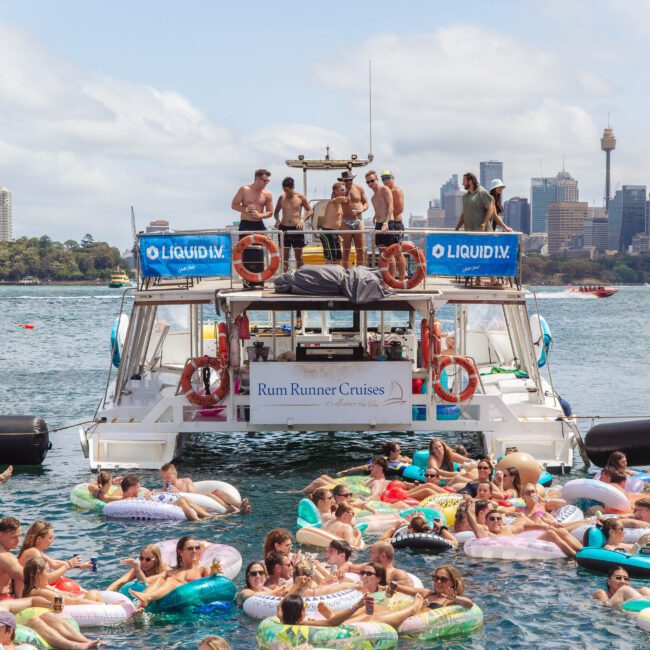 A large group of people on inflatable floats gather around a party boat on the water, with others on the boat deck. The Sydney skyline and Opera House are visible in the background under a partly cloudy sky.