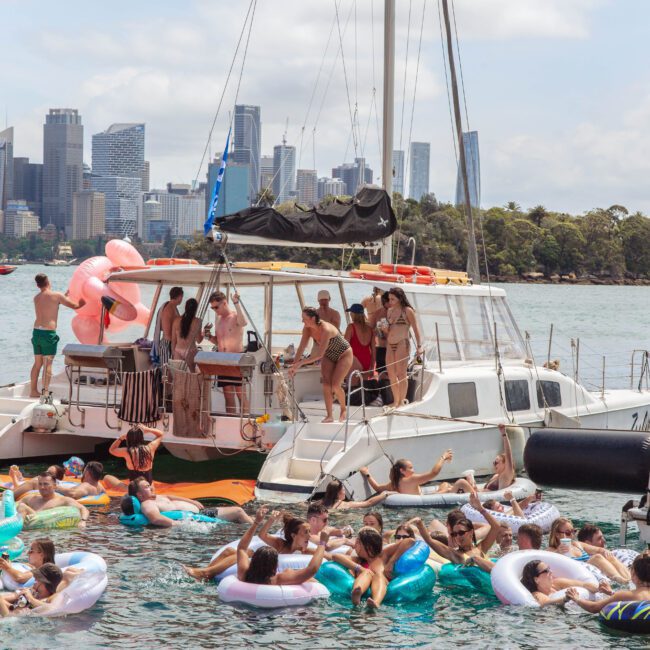 A large group of people on inflatable pool floats gather around a docked catamaran, partying on the water with a city skyline and greenery visible in the background under a sunny sky.