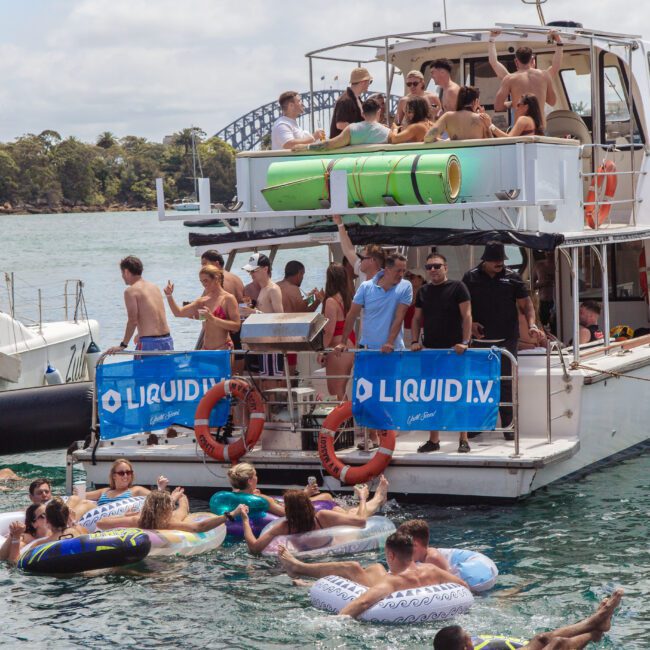 A group of people enjoy a lively party on a docked boat and in the water, surrounded by floaties, with banners reading "LIQUID I.V." visible on the boat. The Sydney Harbour Bridge is in the background.