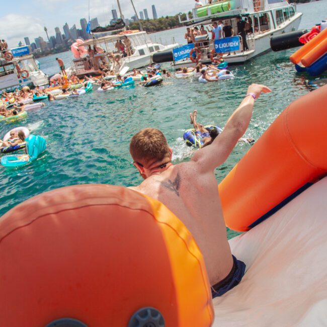 A man slides down an inflatable orange slide into a crowded harbor, where people relax on floaties and boats. The city skyline is visible in the background under a clear sky.