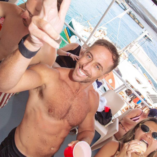 A smiling shirtless man flashes a peace sign and holds a red cup on a boat, surrounded by people enjoying food and drinks near clear blue water and docked boats.