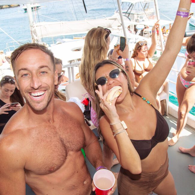 A man and woman in swimsuits smile and pose at a lively boat party. The woman holds a drink and raises her arm, surrounded by people in swimwear, with turquoise ocean and boats in the background.