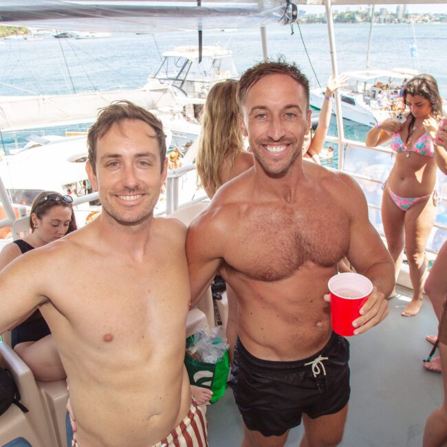 Two smiling men in swimwear pose with red cups at a lively yacht party. Other people in swimsuits are in the background, with boats and water visible outside. The atmosphere is sunny and festive.