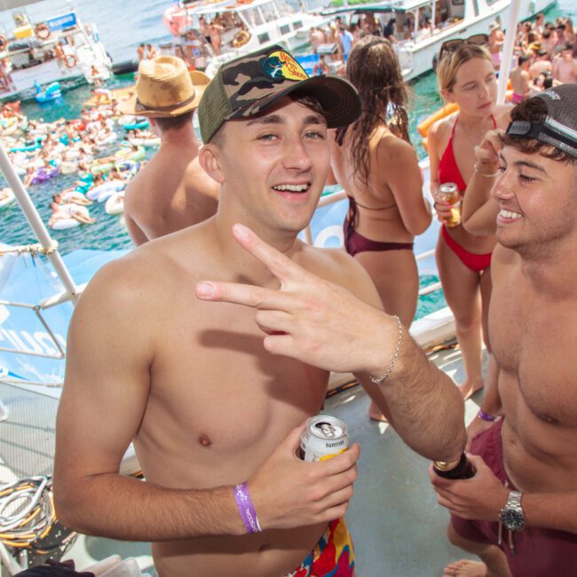 Two young men in swimsuits smile and hold drinks on a crowded party boat, surrounded by other people in swimwear. One man makes a peace sign. Other boats and people on inflatables are visible in the water nearby.