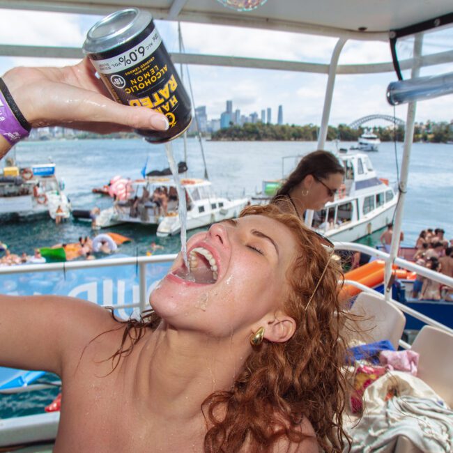 A woman on a boat party tilts her head back, pouring a canned drink into her mouth. Other boats and people are visible in the background, with a city skyline and blue water under a sunny sky.