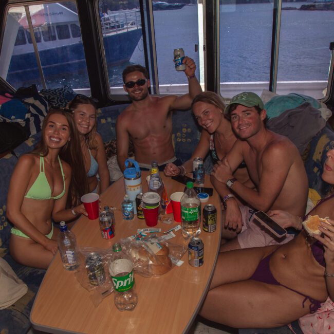 Six young adults in swimsuits sit around a table with drinks and snacks on a boat, smiling and posing for the camera. The boat’s interior and a view of water and another boat are visible through the windows.