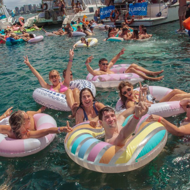 A group of people floats on colorful inflatable rings in clear blue water, smiling and making peace signs. Other partygoers on floats and boats are visible in the background under a sunny sky.