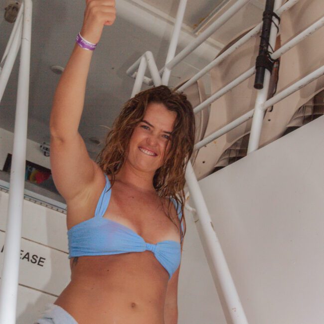A woman in a blue bikini top and towel smiles and points upward while standing on a boat near a staircase. The setting appears sunny and relaxed.