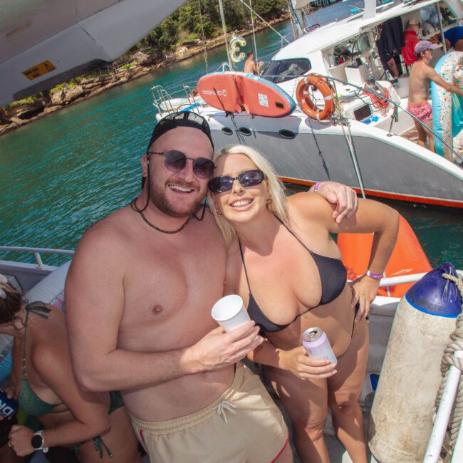 A man and woman in swimsuits smile and pose for a photo on a boat, each holding a drink. Other people are relaxing in the background near clear blue water and another boat. It is a sunny day and everyone appears to be having fun.