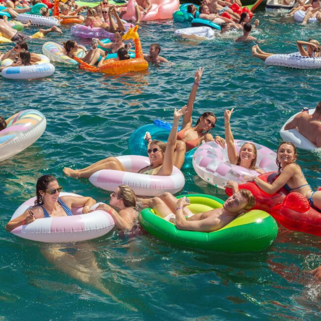 A large group of people relax on colorful inflatable pool floats in the water, enjoying a sunny day. Many are smiling, laughing, and waving, creating a lively, festive atmosphere.