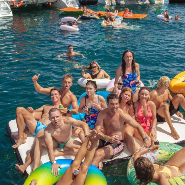 A group of people in swimsuits relax and pose on large floating mats and inner tubes in bright blue water, surrounded by others swimming and lounging on floats near anchored boats.