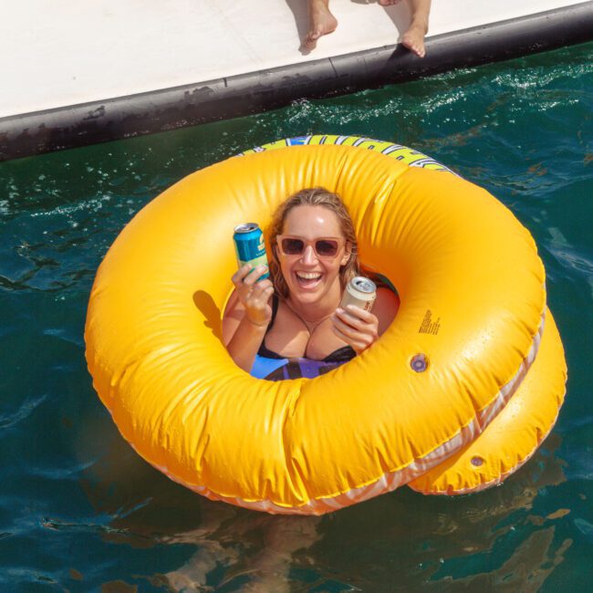 A smiling woman floats on turquoise water in a large yellow inflatable ring, holding two canned drinks, with part of a boat and another person’s feet visible above her.