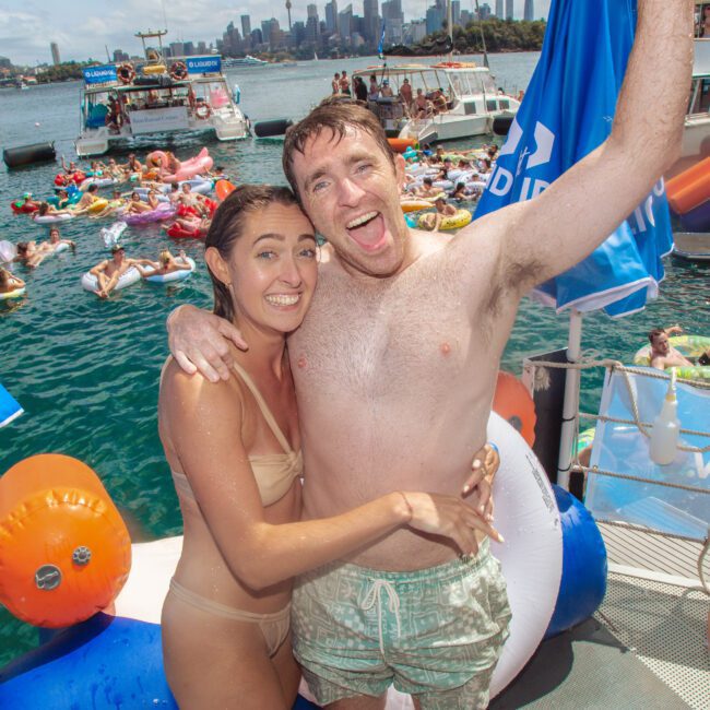 A smiling man and woman in swimsuits hug on a docked boat at a lively water party, with people swimming and lounging on inflatables in the water and other boats nearby, city skyline in the background.