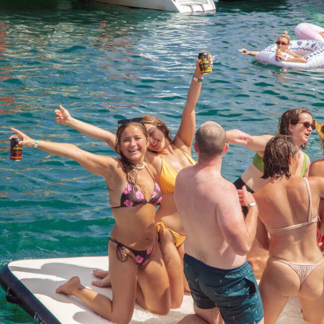 A group of people in swimsuits celebrate on a floating platform in blue water. Some hold drinks and smile at the camera, while others talk and relax. More people float on inflatables in the background.