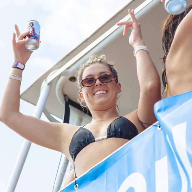 A woman in sunglasses and a black bikini holds up a can and raises her arms, smiling on a boat next to another woman. A blue banner and a partly cloudy sky are visible in the background.
