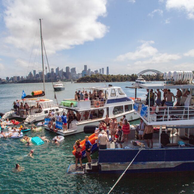People gather on boats and float on inflatables in a lively party scene on the water, with Sydney’s city skyline and the Harbour Bridge visible in the background under a partly cloudy sky.