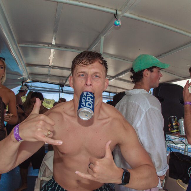 A shirtless man on a boat holds a Carlton Dry can in his mouth and makes a hand gesture, surrounded by people drinking and socializing. Sunlight streams in and the mood is festive.