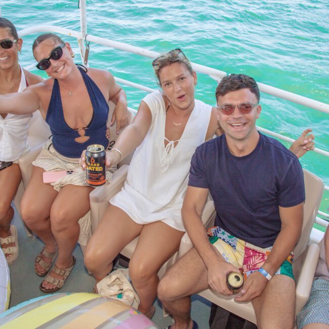 Four people are sitting on a boat, smiling and holding drinks, with bright sunlight and turquoise water in the background. Inflatable pool floats are visible in the foreground.