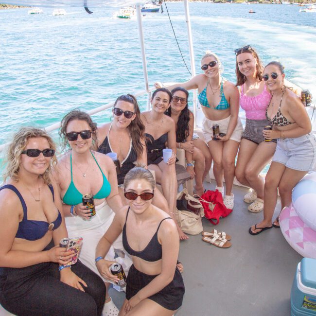 A group of nine women in swimsuits and sunglasses smile and pose together on a boat with water and other boats in the background. An inflatable pool ring and cooler are visible on the deck.