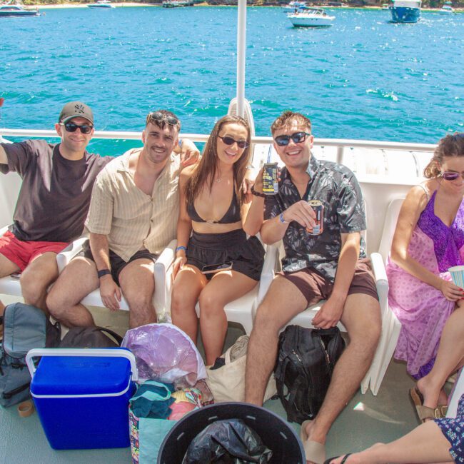 Five people sit and smile on a boat, holding drinks, with blue water and boats in the background. They wear summer clothes and sunglasses, enjoying a sunny day. A cooler and bags sit at their feet.