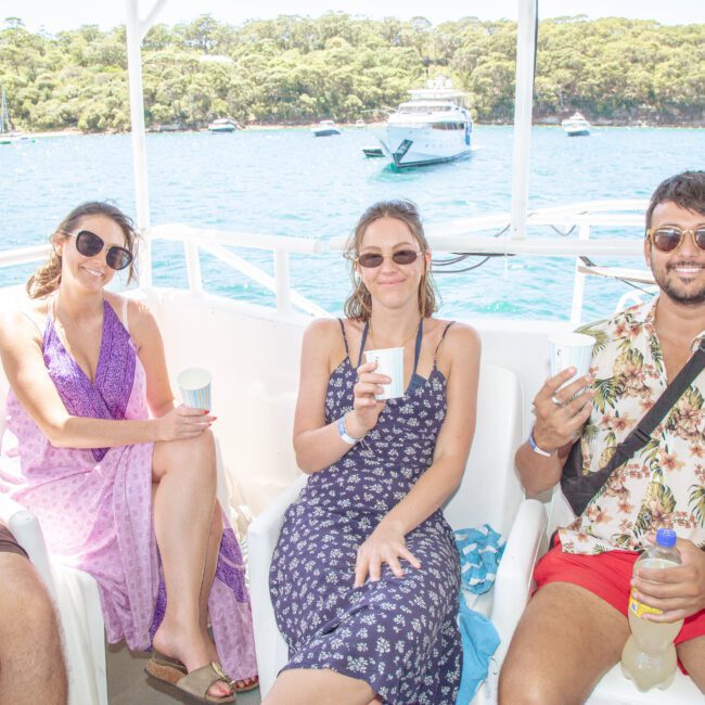 Three people sit on a boat, smiling and holding drinks. The background shows blue water, a yacht, and a forested shoreline under a sunny sky.