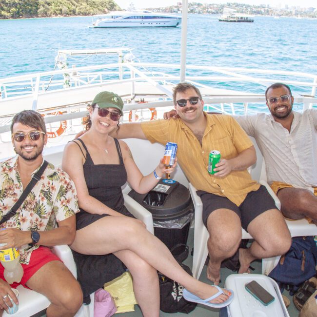 Four people sit on white chairs on a boat, smiling and holding up drinks. The blue ocean and other boats are visible in the background under a sunny sky. They appear relaxed and are wearing summer clothes.