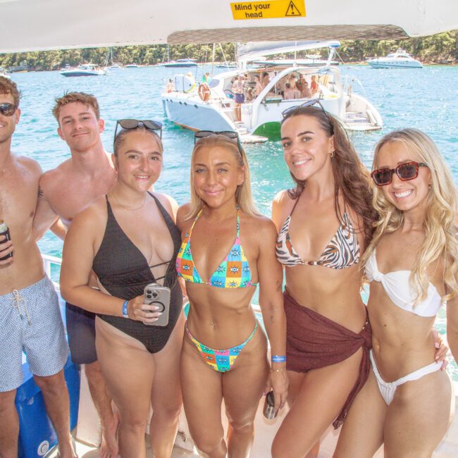 Six young adults in swimsuits smile and pose together on a boat in sunny weather, with boats and turquoise water in the background, suggesting a fun day out on the water.
