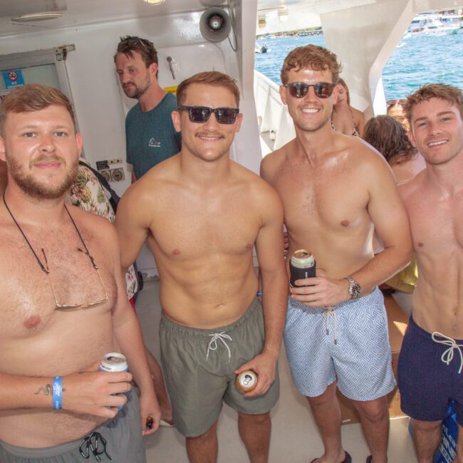 Four smiling men in swim trunks stand together on a boat, holding drinks. The boat is crowded with people enjoying sunny weather, and the sea with a coastal view is visible in the background.
