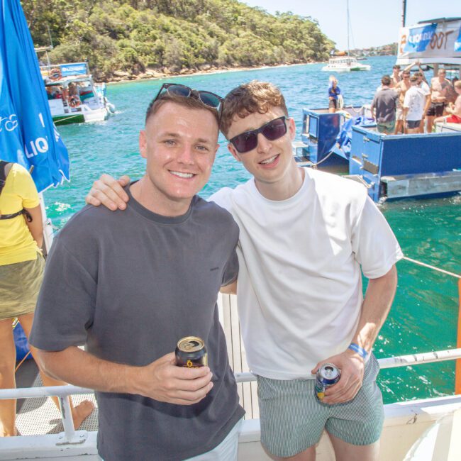 Two young men smile and pose with drinks on a boat. Other people and boats are visible nearby on blue-green water, with a lush green shoreline in the background. It is a sunny day, and the scene appears relaxed and festive.