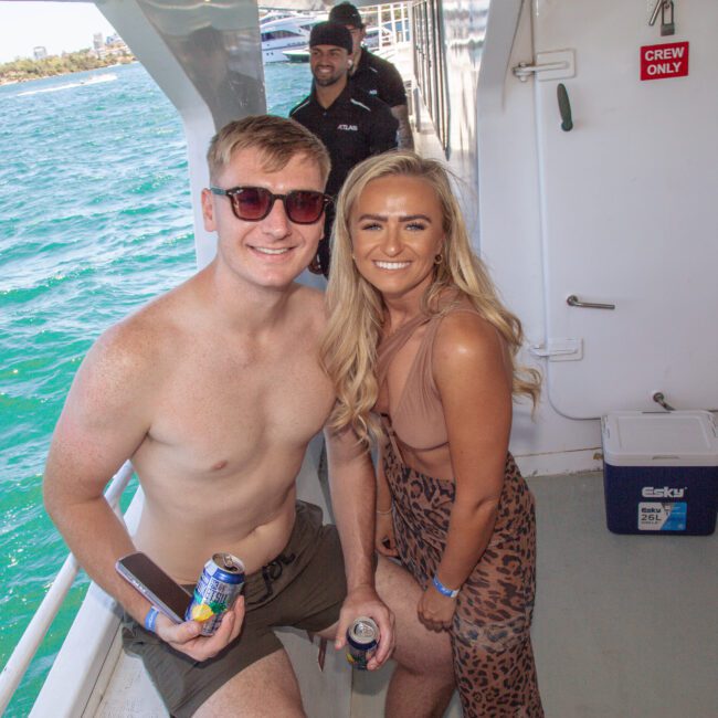 A smiling man in swim shorts and sunglasses stands next to a smiling woman in a tan crop top and skirt on a boat by the water. Another man is visible in the background. Coolers and a "Crew Only" sign are nearby.