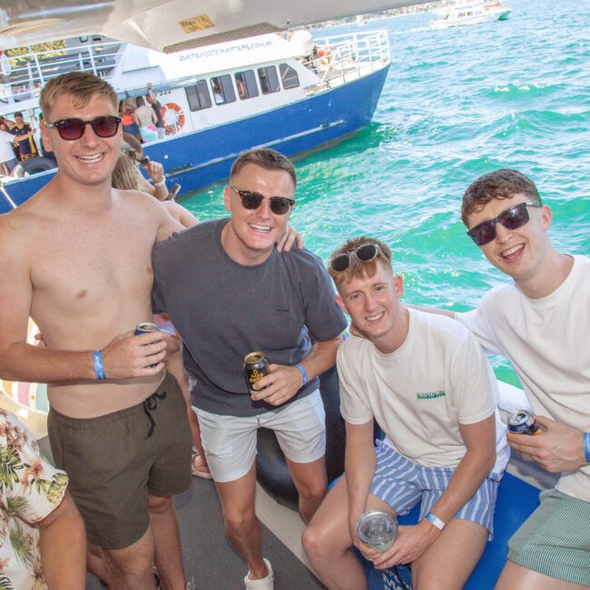 Four young men wearing sunglasses and casual summer clothes smile while sitting on a boat with drinks in hand; turquoise water and another boat with people are visible in the background.