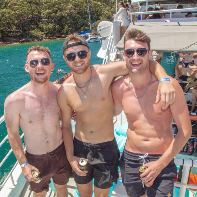 Three young men wearing sunglasses and swim trunks stand arm-in-arm, smiling on the deck of a boat in sunny weather. They are holding drinks, with water and other people in swimwear visible in the background.