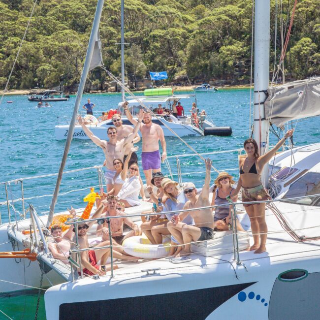 A group of people in swimwear enjoy a sunny day on a catamaran, waving and smiling at the camera. The boat is on blue water near a forested shoreline, with other boats visible in the background.