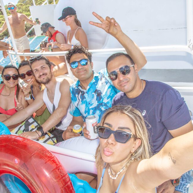 A group of friends in swimsuits smile and pose for a selfie on a sunny boat deck, holding drinks and pool floats. The background shows more people relaxing near railing and blue water.