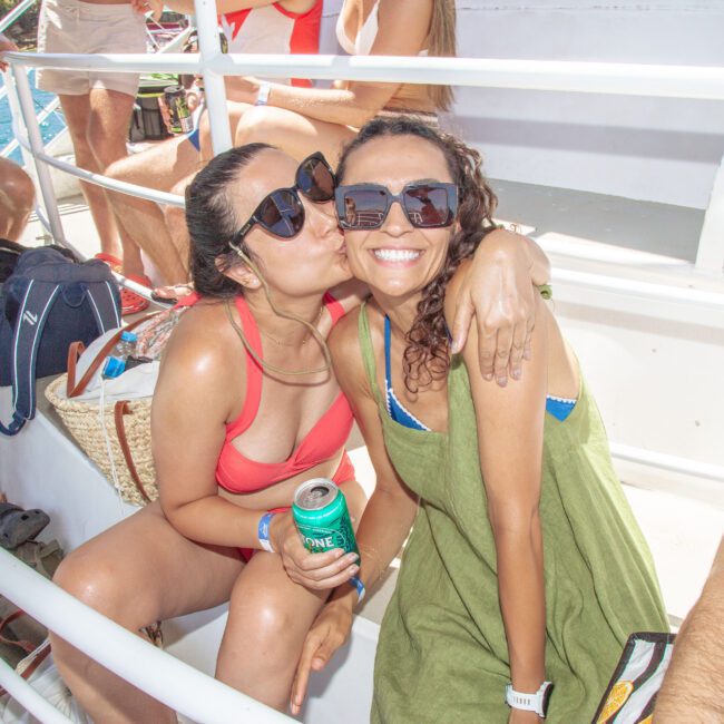 Two women smiling on a boat; one in a red bikini top kisses the other, who wears a green dress. Both wear sunglasses, surrounded by other people, bags, and drinks, enjoying a sunny day outdoors.