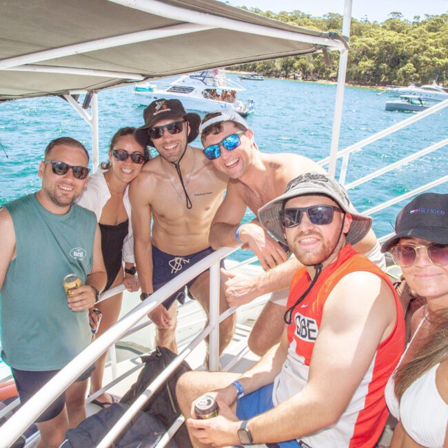 A group of six smiling people in casual summer clothes and sunglasses pose together on a boat in sunny weather, with blue water and other boats visible in the background.