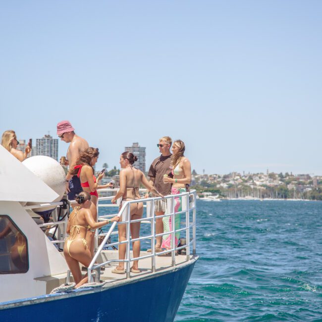 A group of people in swimwear stand and relax on the deck of a boat on a sunny day, with a city skyline and waterfront visible in the background.