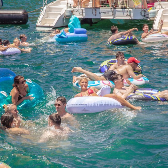 A group of people float on inflatables and swim in bright blue water near a docked boat during a sunny day, enjoying a lively social gathering.