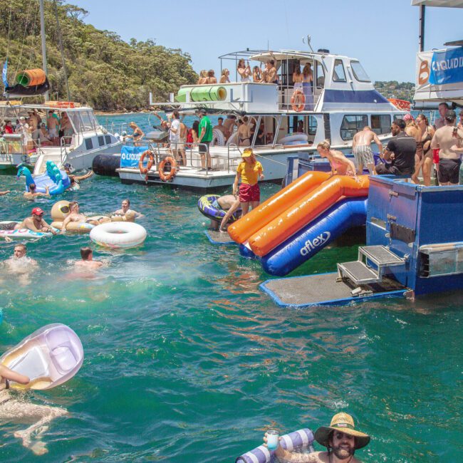 A lively boat party with people swimming and lounging on inflatables in clear blue water. Multiple boats are anchored, with slides leading into the water and groups socializing on decks under sunny skies.