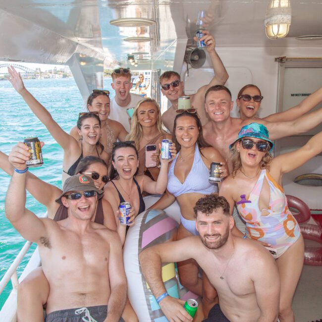 A group of young adults in swimsuits smile and hold drinks while posing together on a boat, with blue water and a distant bridge visible in the background. The atmosphere is lively and festive.