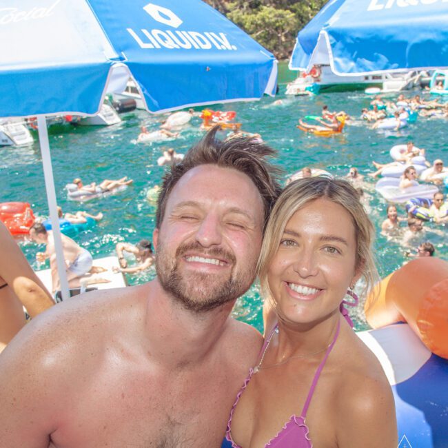 A man and a woman in swimsuits smile at the camera at a lively pool party, surrounded by people on inflatables in the water, with blue umbrellas and trees in the background.