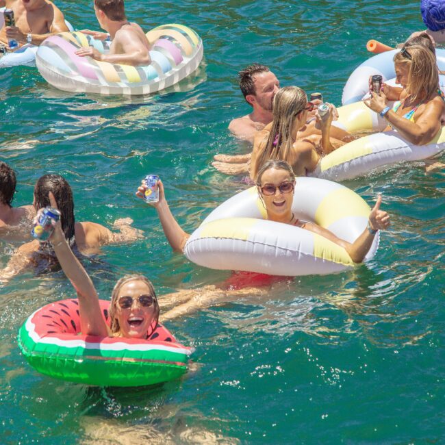 A group of people relax and have fun on colorful pool floats and in clear blue water, smiling and holding drinks under the sun. The atmosphere is festive and lively. In the corner, a logo reads “Yacht Social Club.”.