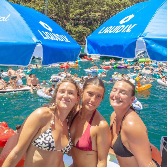 Three women in bikinis smile under blue umbrellas on a boat, with a crowded lake full of people on colorful inflatables and green trees in the background.