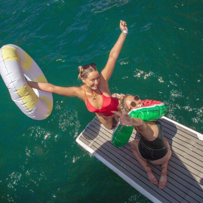 Two women in swimsuits stand on a dock beside water, smiling and holding pool floats. One woman wears a red suit and yellow-and-white float; the other wears black and holds a watermelon float. Both appear joyful and excited.