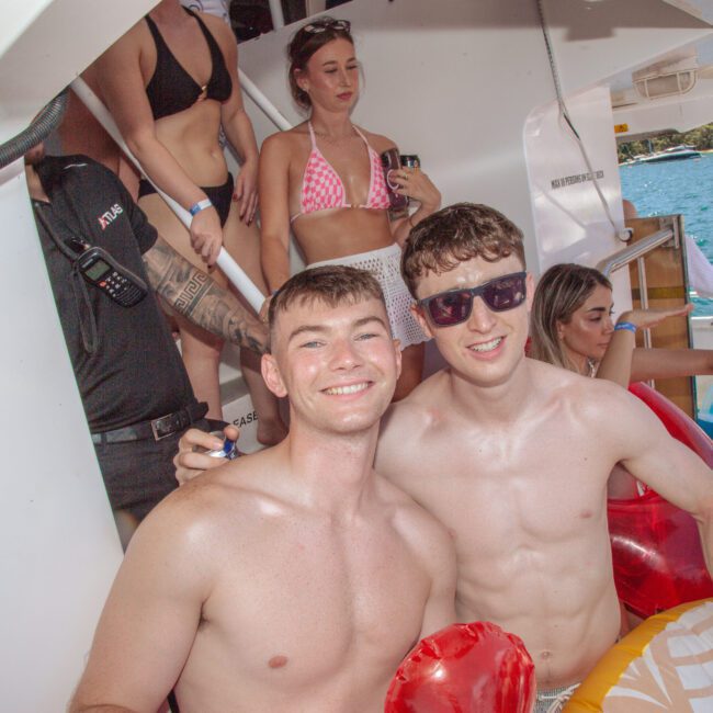 Two young men in swim trunks smile at the camera on a boat, holding inflatable pool rings. Several other people in swimwear are in the background, enjoying the sunny day by the water.