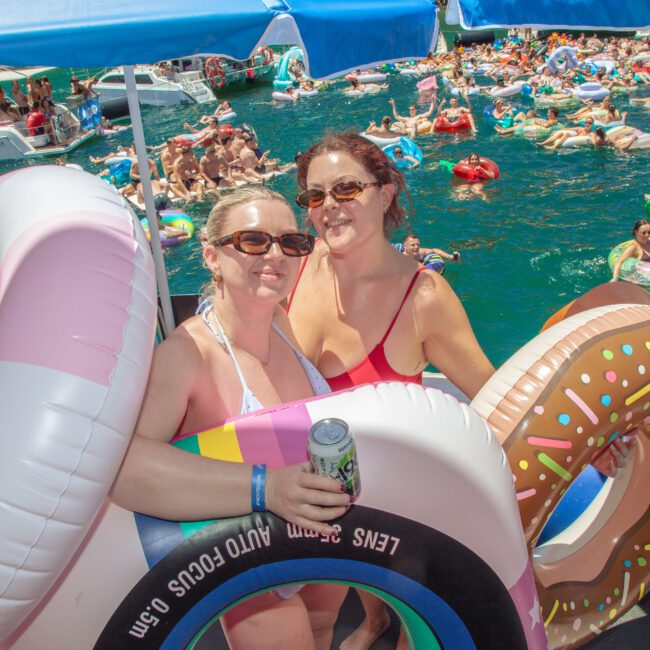 Two women in swimsuits smile on a boat, each holding colorful inflatable pool floats shaped like donuts. A crowd of people on floats relaxes in the water behind them under bright sunlight.