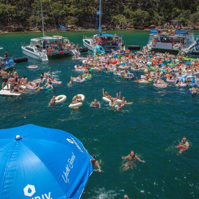A large group of people in swimsuits float on inflatable rings and lounge in the water near several boats, with blue umbrellas in the foreground and trees lining the shore in the background.