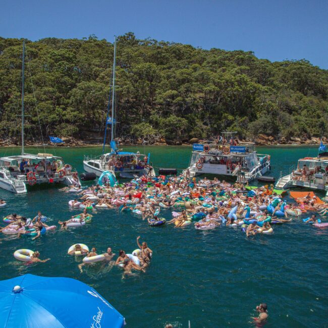 A large group of people relax on colorful inflatables in the water, surrounded by several anchored boats near a forested coastline on a sunny day. A blue umbrella and a "Yacht Social" logo are visible in the foreground.