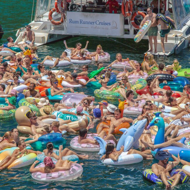 A large group of people relax on colorful inflatable floaties in the water near a docked Rum Runner Cruises boat on a sunny day. The scene is lively, with many adults enjoying the summer event.